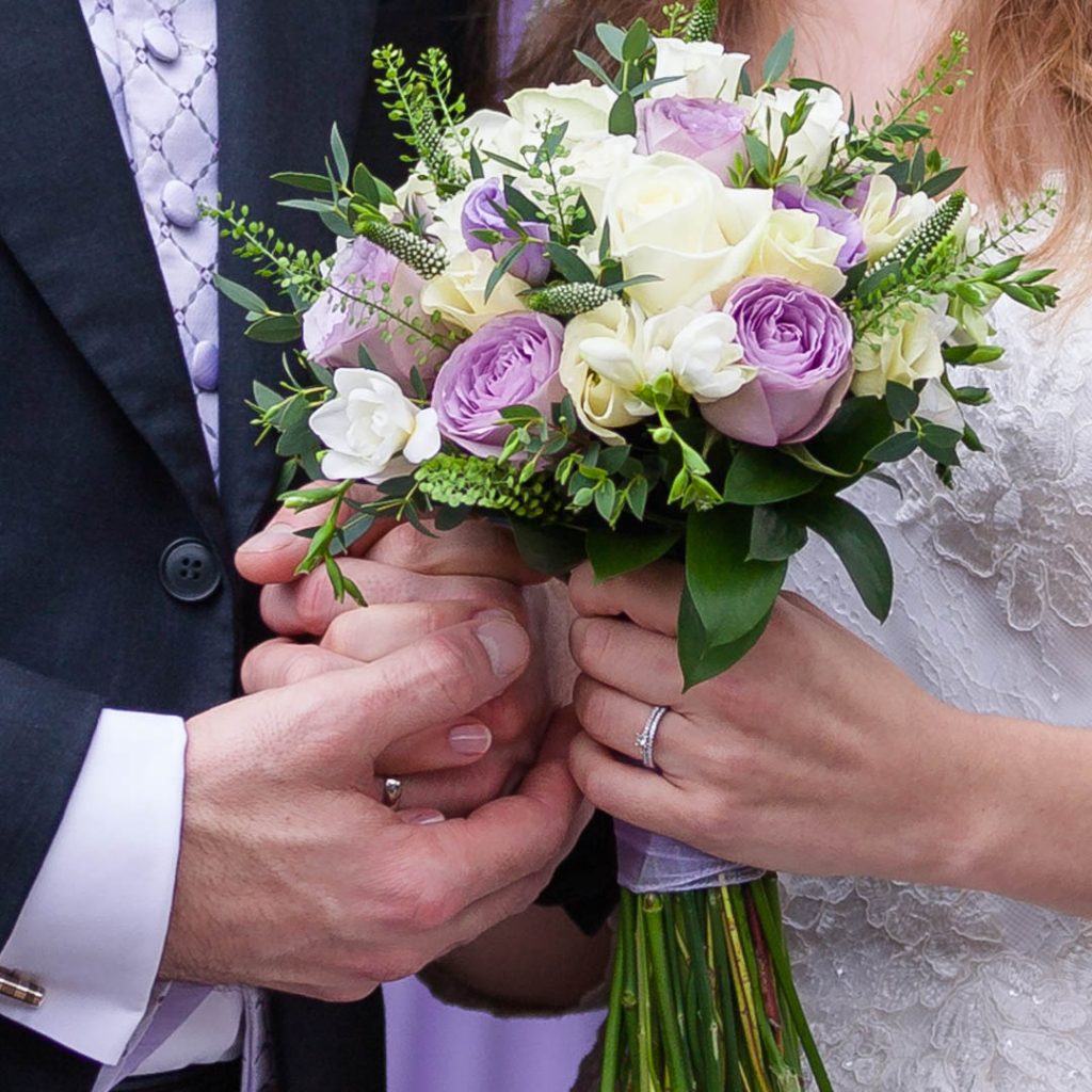 Hands of bride and groom entwine around her bouquet