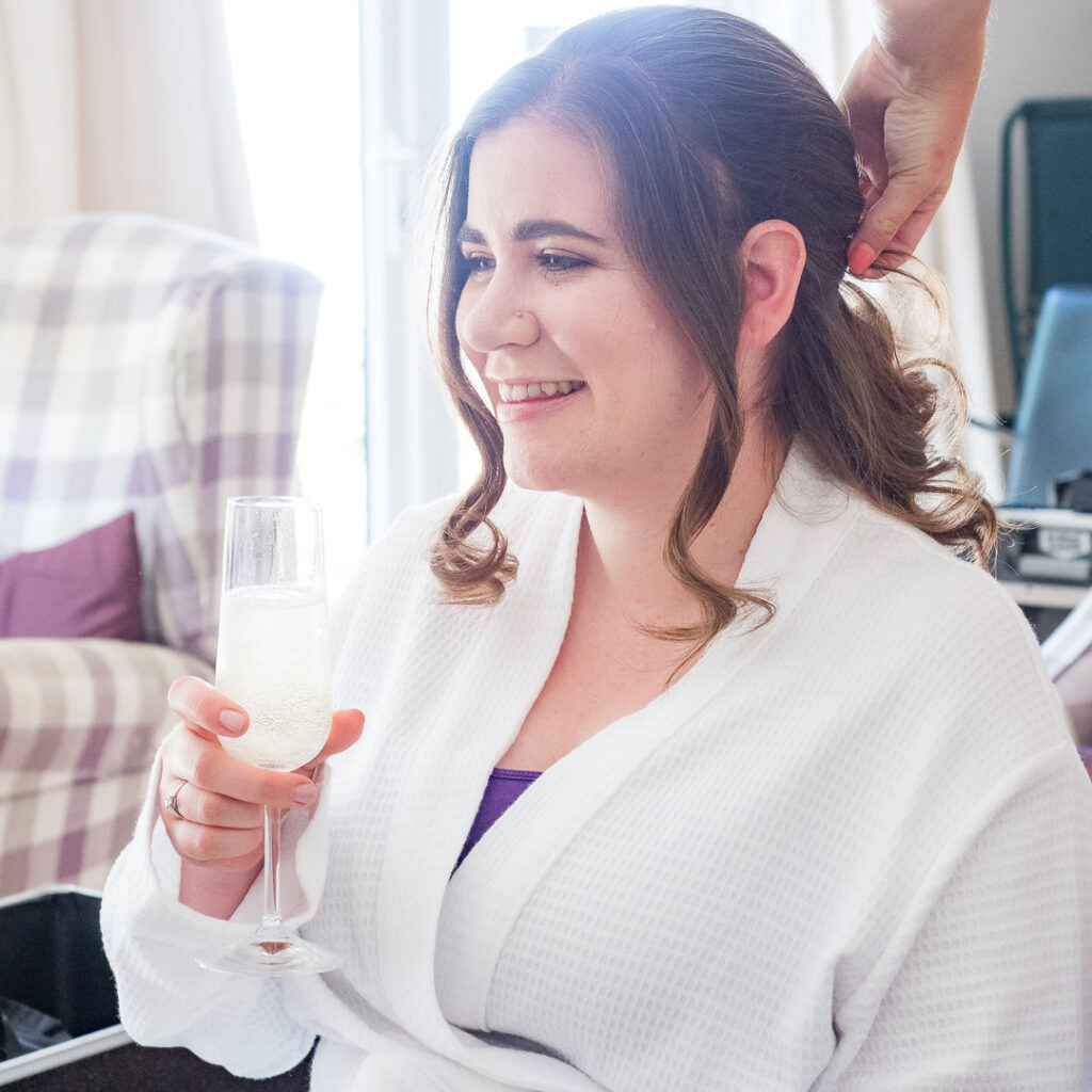 Bride enjoys glass of prosecco while her har is styled for her wedding