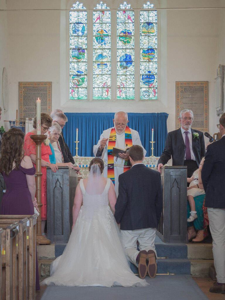 Vicar blesses couple kneeling before altar durng classic English country church wedding