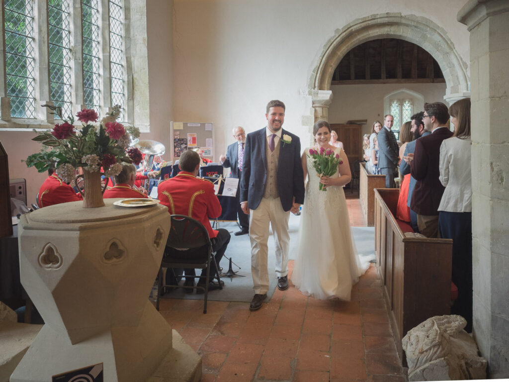 Bride and groom walk down aisle at end of  classic English country church wedding service as brass band plays
