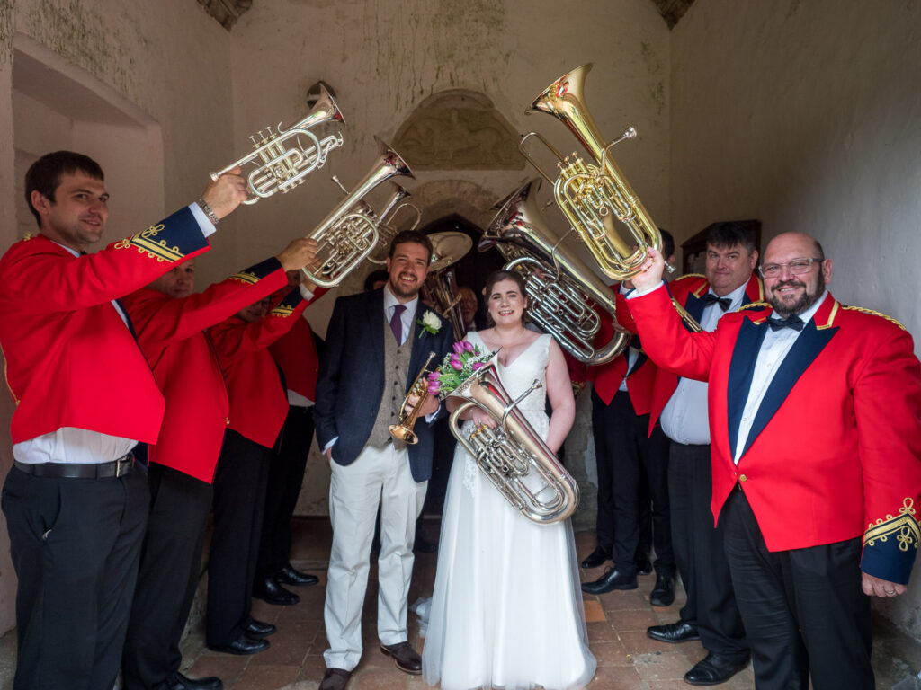 Brass band in red jackets form guard of honour for bride and groom in English country church porch