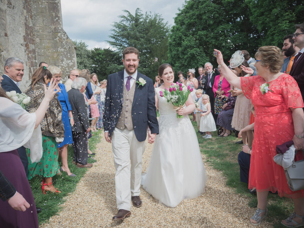 Guests throw confetti over bride and groom in churchyard  after classic English country church wedding