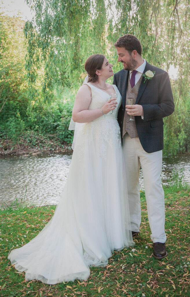 Romantic portrait of bride and groom by stream lit by sun shafting through weeping willow
