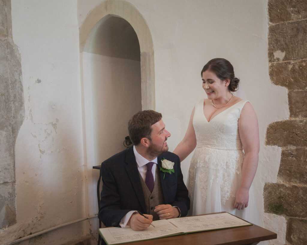 Standing bride smiles at seated groom singing register classic English country church wedding