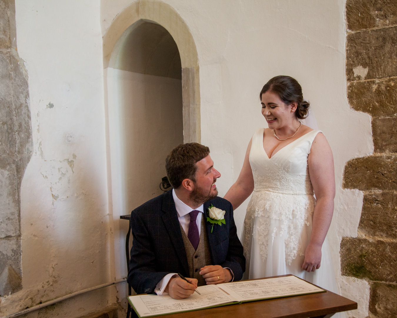 Standing bride smiles at seated groom singing register classic English country church wedding