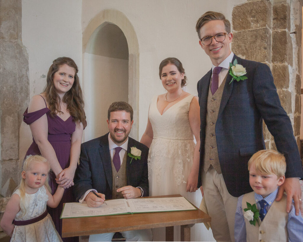 Seated groom with bride, bridesmaids, best man and marriage register during classic English country church wedding