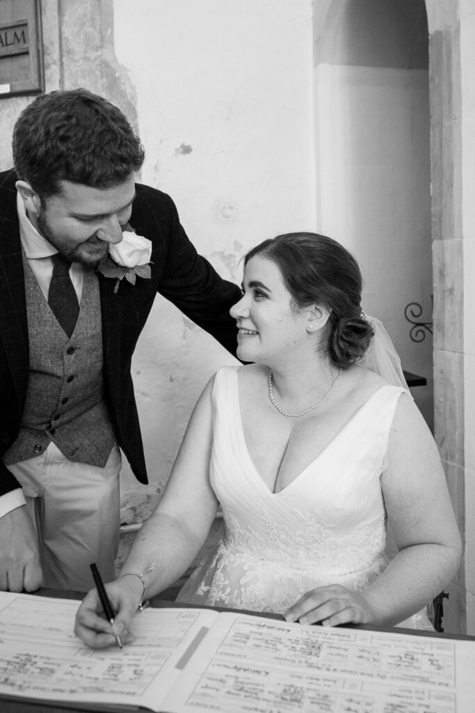 Groom looks on as bride signs register during classic English country church wedding