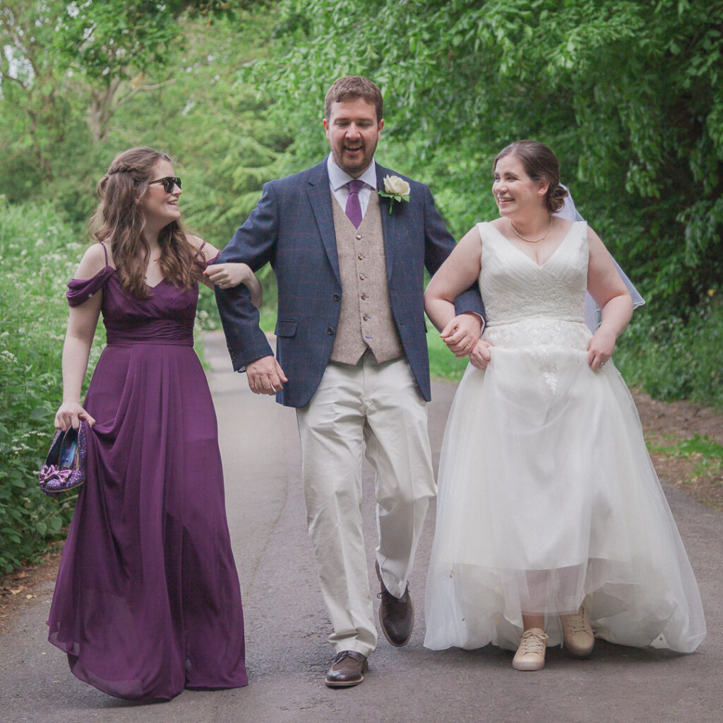 Bride, groom and bridesmaid walk together down English country lane after wedding
