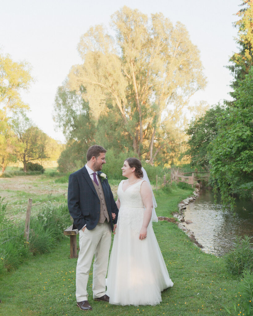 Bride and groom smile at each other in romantic portrait beside country stream