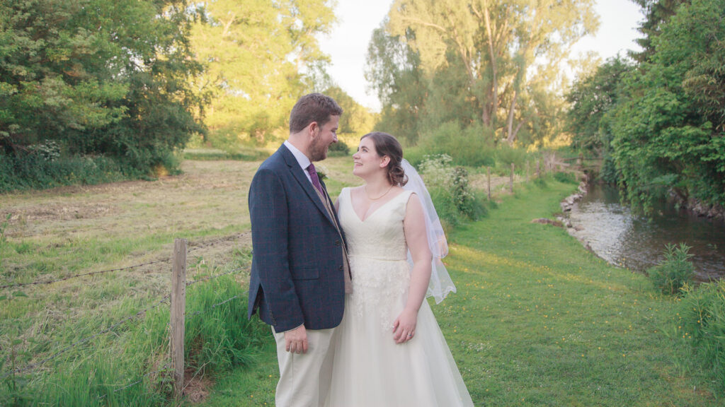 Bride and groom smile at each other in romantic portrait beside country stream