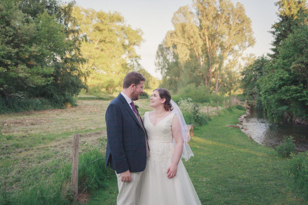 Bride and groom smile at each other in romantic portrait beside country stream