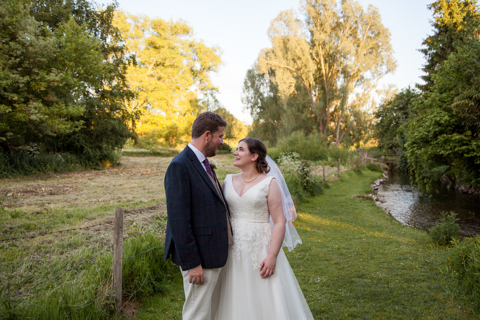 Bride and groom smile at each other in romantic portrait beside country stream