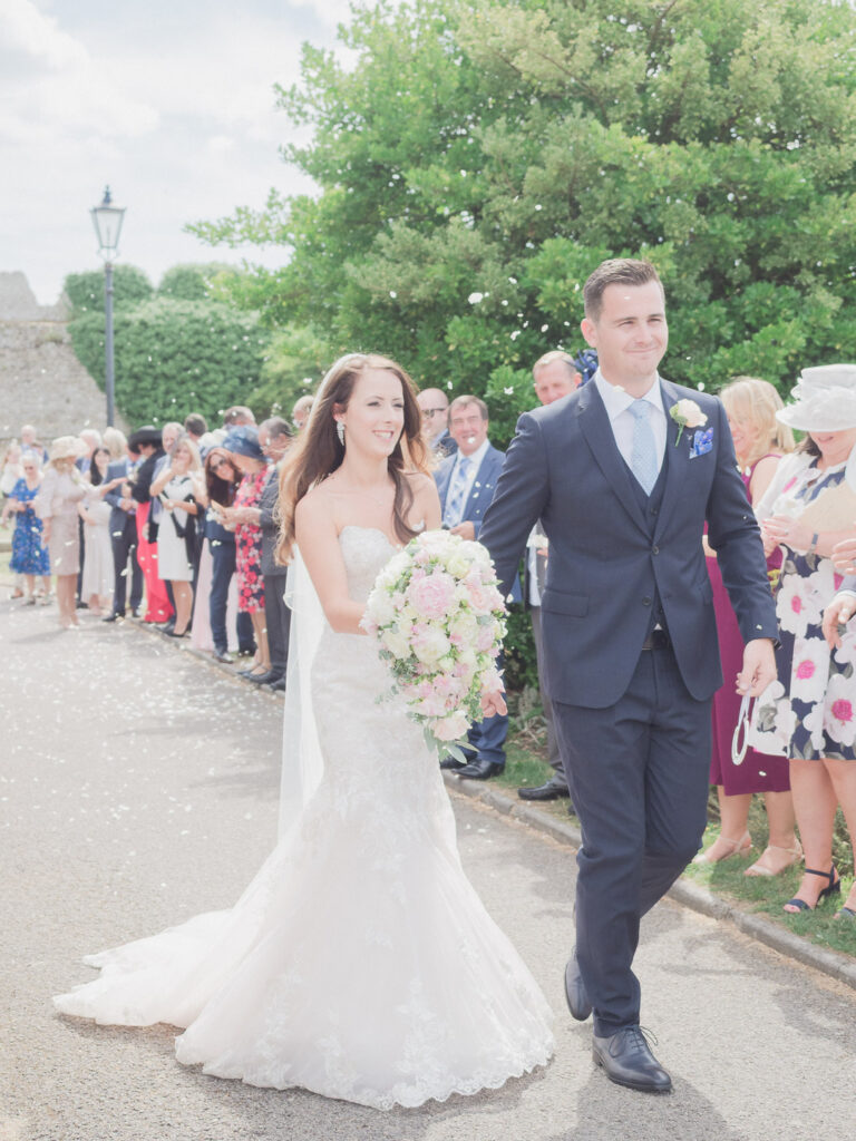 Holding her bouquet, bride walks with groom past wedding guests throwing confetti