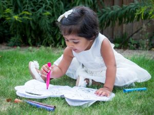Indian girl toddler in white dress with colouring book sitting on lawn