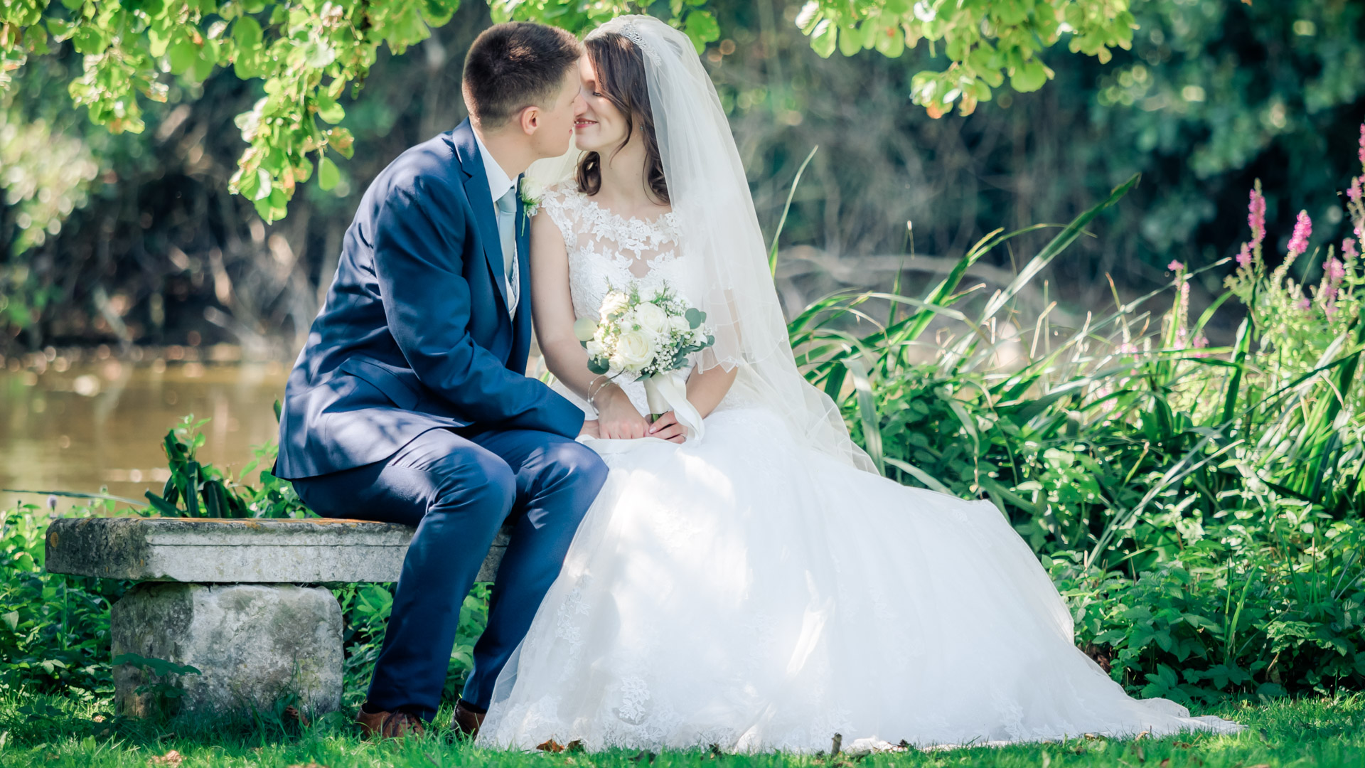 Bride and groom enjoy romantic moment seated on a stone bench in front of a lake in summertime