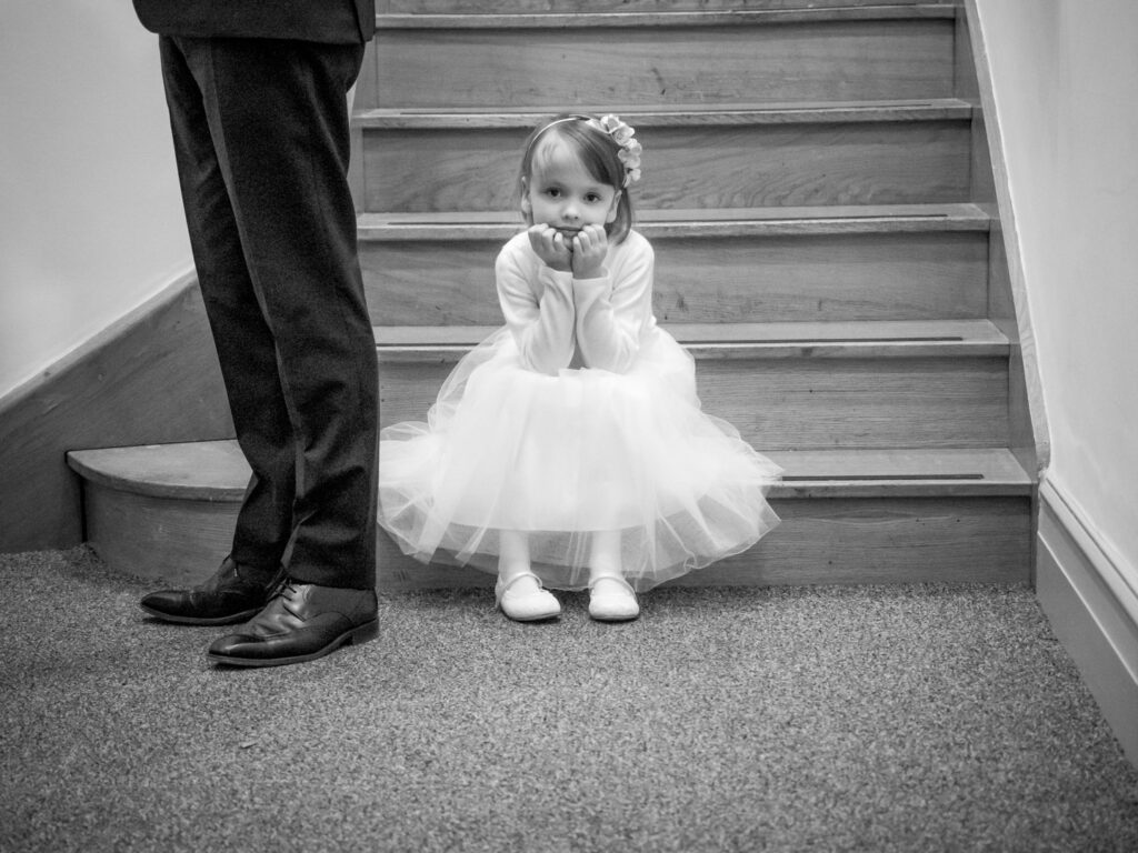 Little bridesmaid sits on a bottom step with her father's legs in shot giving a sense of scale