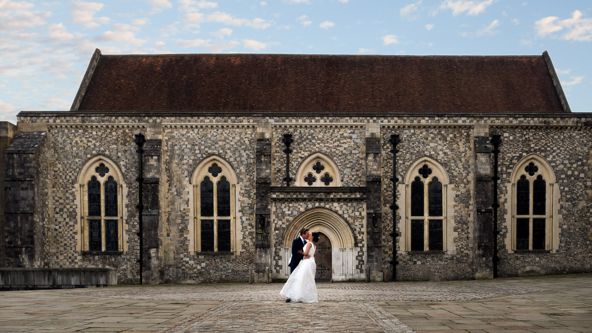 Groom kisses bride outside Winchester Great Hall beneath bue sky at sunset