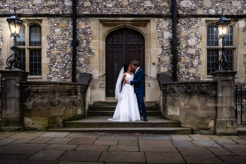 Bride and groom kiss on steps of Victoran Gothic flint-faced building lit by wrought-iron lamps