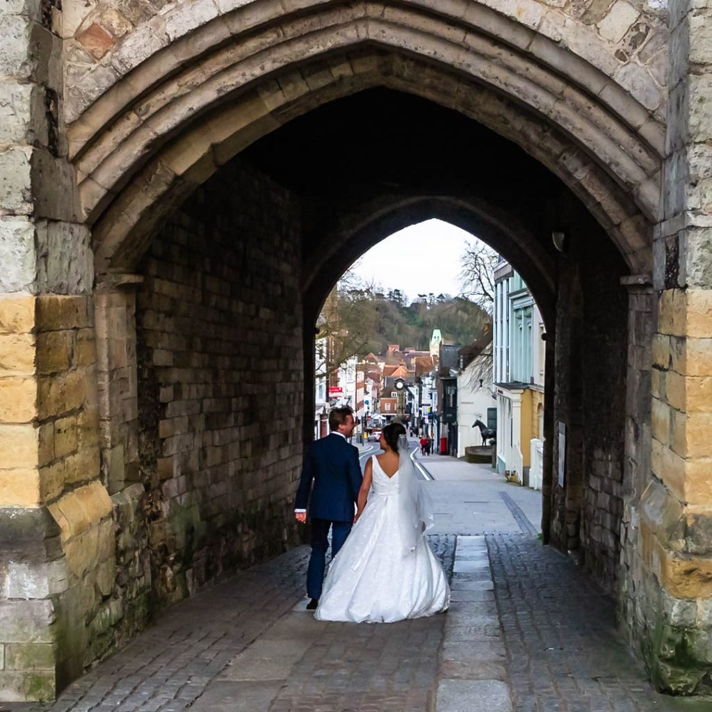 Bride and groom walk through the arch of Winchester's Westgate down towards the city centre