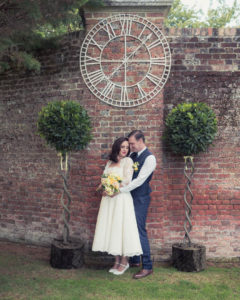 Groom holds bride beneath a sundial flanked by ornamental bay trees at The Orangery Suite
