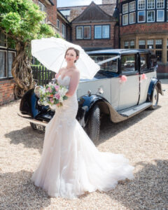 Bride holds a sun parasol and bouquet in front of a vintage Rolls Royce at The Montagu Arms Hotel, Beaulieu
