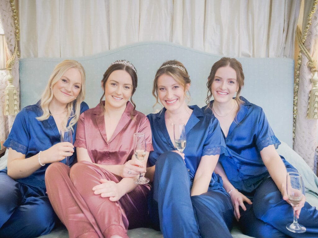 Bride and her bridesmaids in satin pyjamas drinking champagne together on her bed