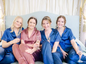 Bride and her bridesmaids in satin pyjamas drinking champagne together on her bed