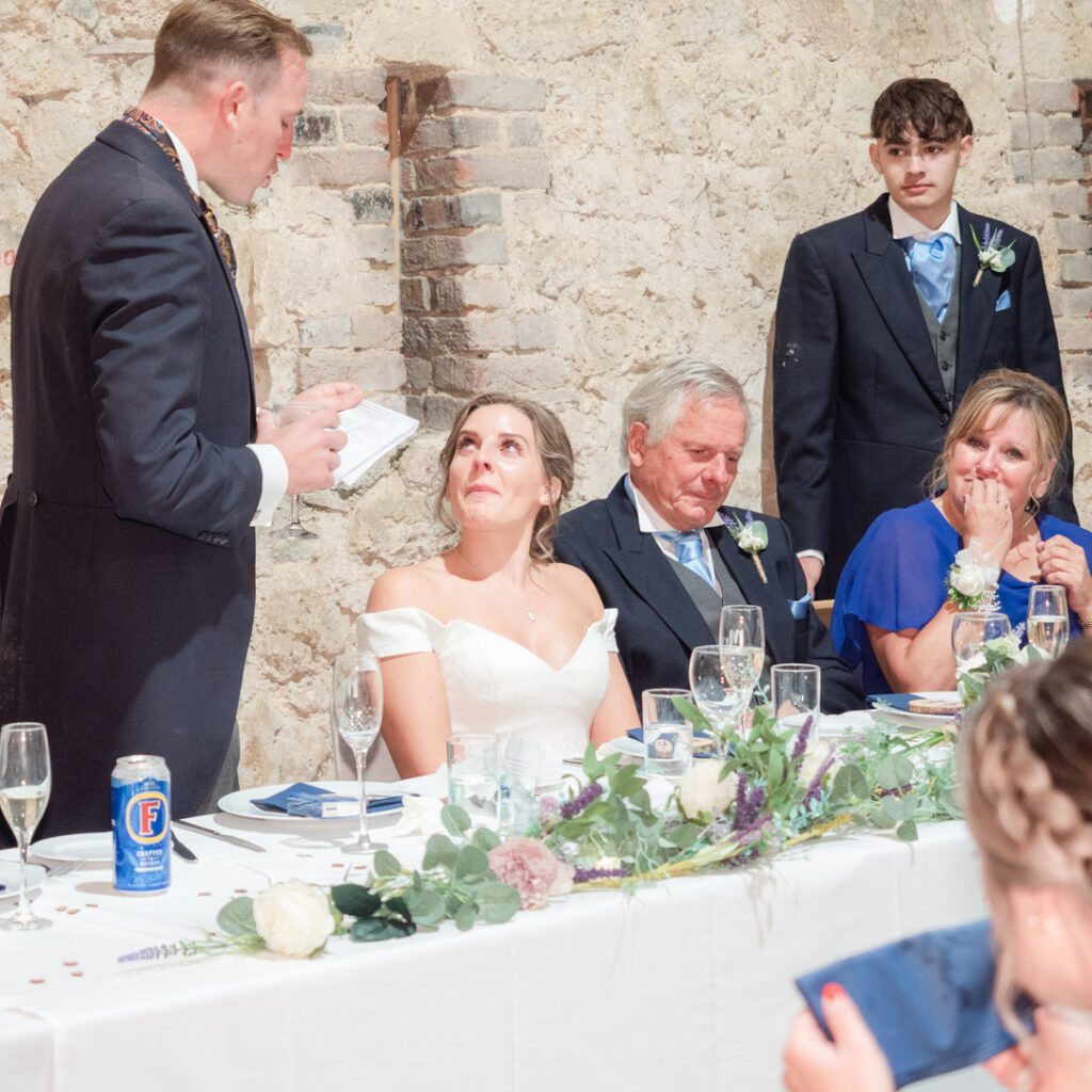 Bride looks tearfully at groom during his speech at their wedding reception in Titchfield Great Barn