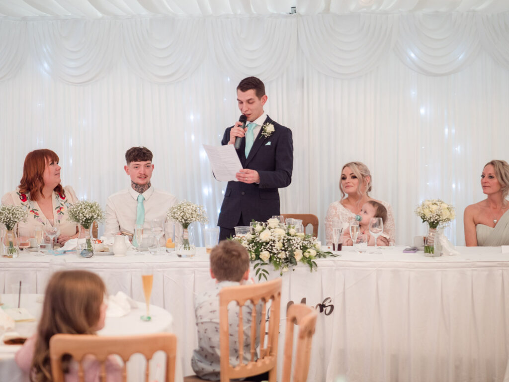 Groom gives speech as his bride and wedding guests look on