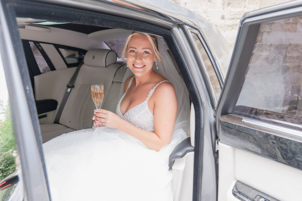 Bride holds champagne flute in wedding car outside Romsey Abbey