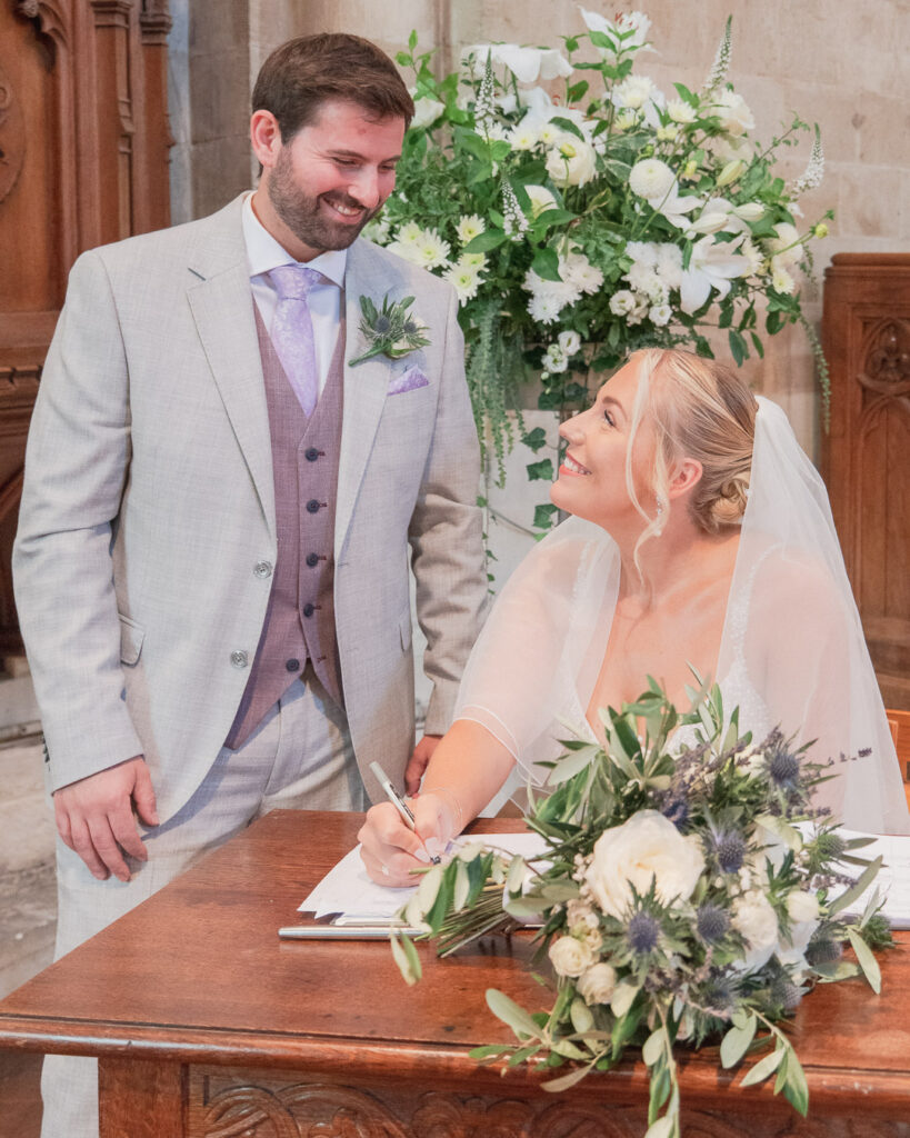 Bride smiles at groom in Romsey Abbey as she signs the register