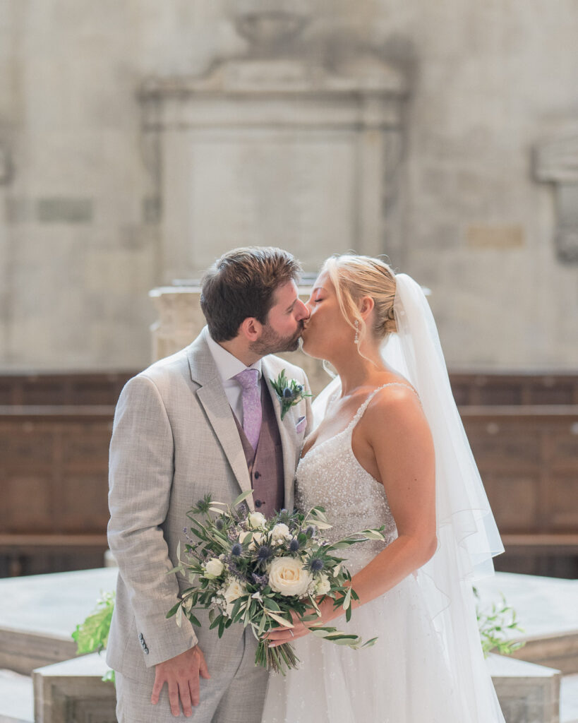 Bride and groom kiss in front of the font in Romsey Abbey